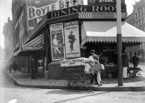 A black and white photograph of a man and boy on a street corner, working with a pushcart.