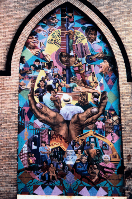 A mural on a building facade. In the center, two hands hold a group of people, looking upwards and at each other. The mural is filled with colors: blues, yellows, purples, pinks, greens.