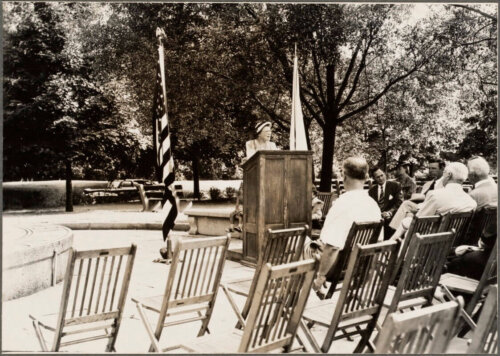 A black and white photograph of a woman standing at a rostrum speaking to a crowd.