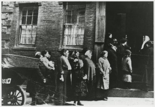 A black and white photograph of women in dresses standing in line outside of a brick building.