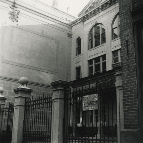 A black and white photograph of a large concrete building with arched windows, a pediment, and a gate out front.