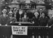A black and white photograph of the inside of a shul, with interior decorations, men standing in suits and hats, and a sign that reads: "built by Sam Katz 1920."