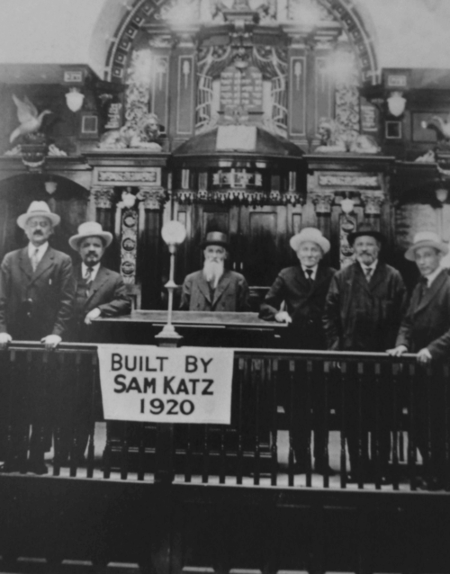 A black and white photograph of the inside of a shul, with interior decorations, men standing in suits and hats, and a sign that reads: "built by Sam Katz 1920."