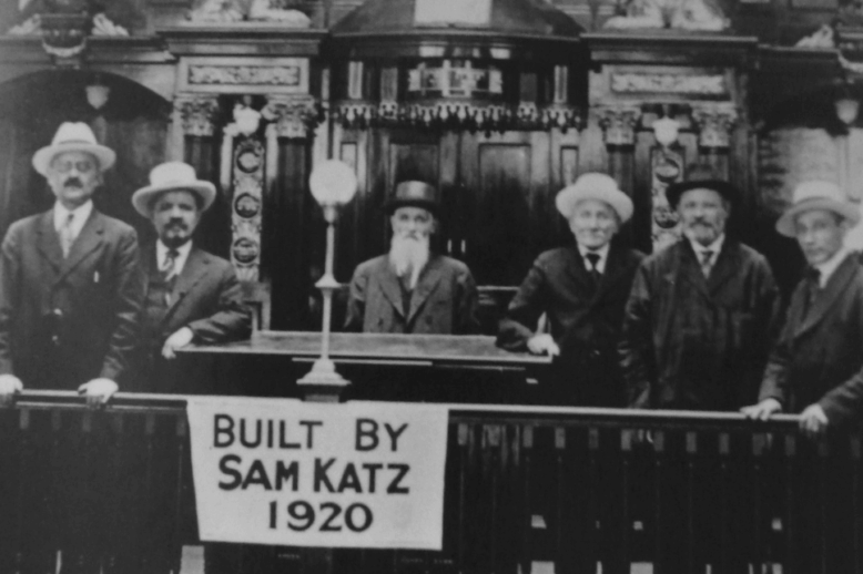 Shul Interior A black and white photograph of the inside of a shul, with interior decorations, men standing in suits and hats, and a sign that reads: "built by Sam Katz 1920."