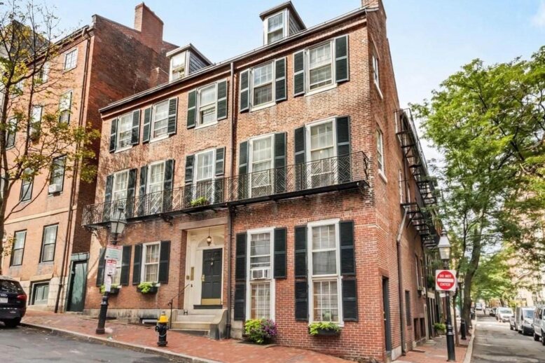 A four story brick building at the corner of a street with a cobbled sidewalk. The building facade has many windows with black shutters.