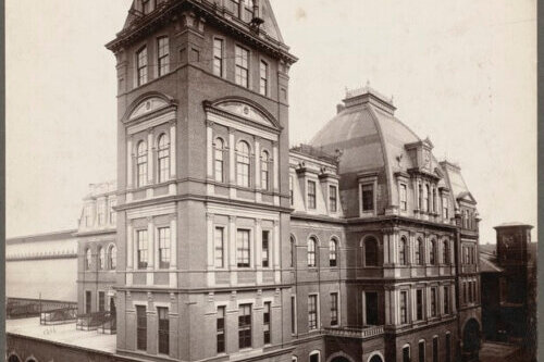 A Second French Empire style building with a tower and domed entry hall. There are many windows with elaborate trim and people and horse-drawn wagons wait outside.