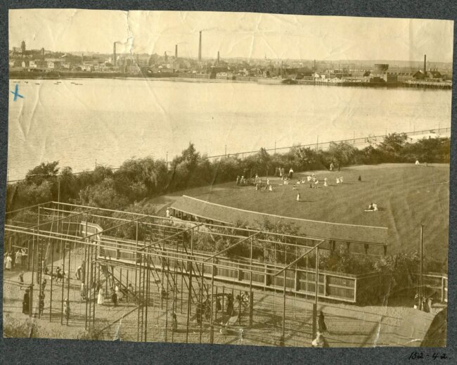 womens_gym(1897)(Olmsted_shurtleff) Sepia image of a gymnasium structure and lawn on the edge of a river.
