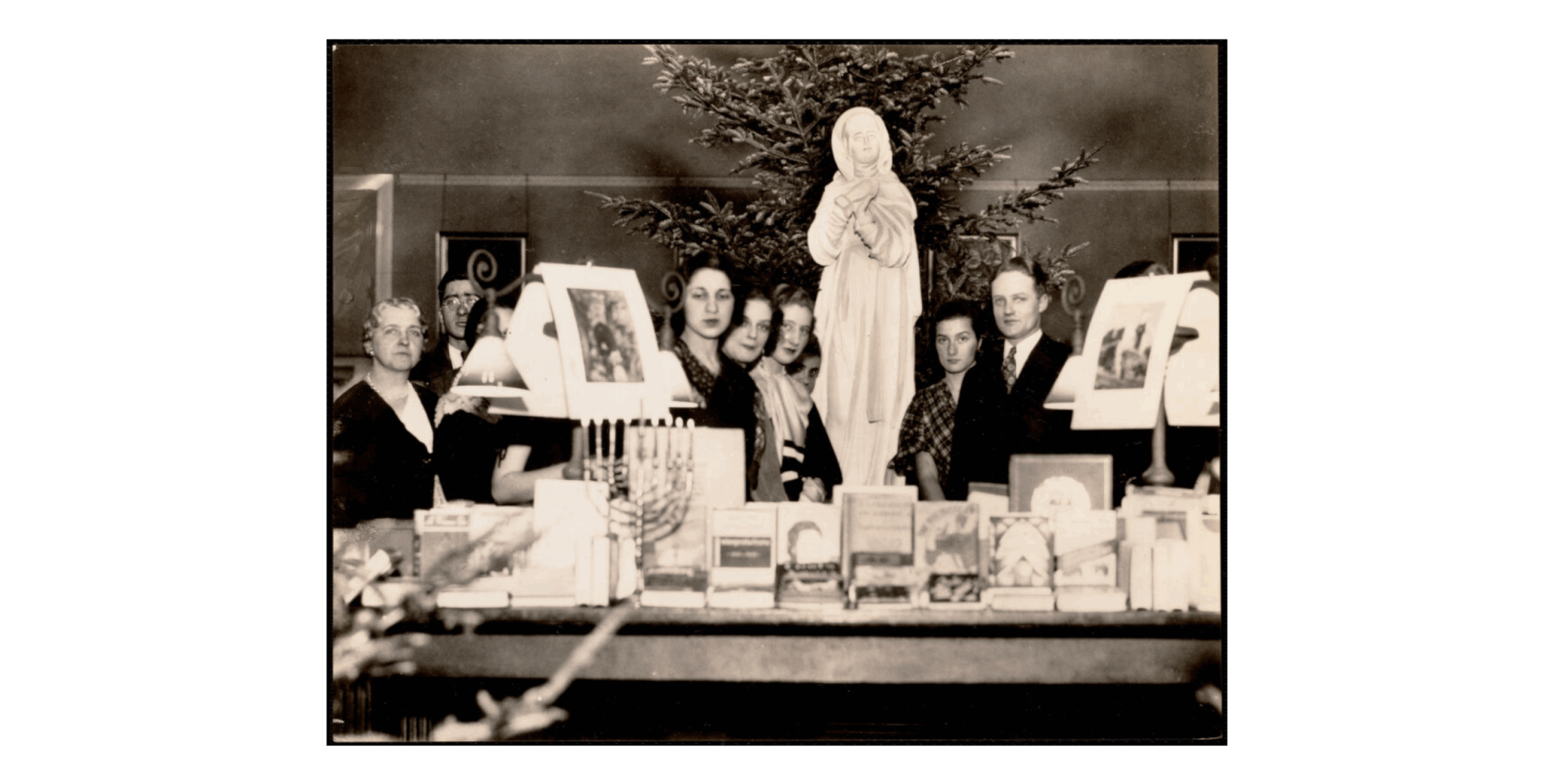 A group of people stand behind a table with Christmas and Hanukkah decorations including a large Virgin Mary Statue.