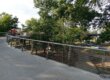 An image of a paved bridge with wire railings overlooking trees.