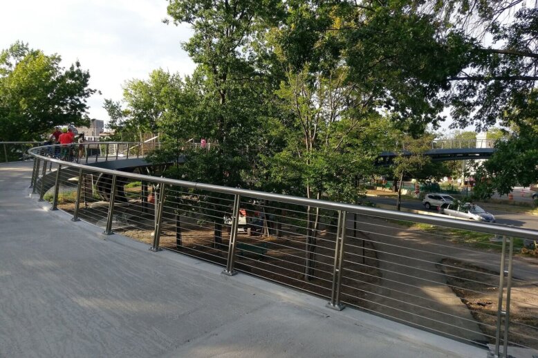 An image of a paved bridge with wire railings overlooking trees.
