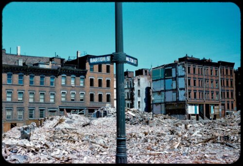 Photograph of a street corner sign in front of a rubble field with tenement houses behind it.
