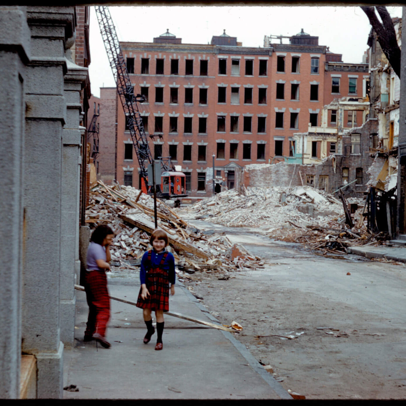 Two children standing next to a concrete building in front of a tall brick building and rubble pile.