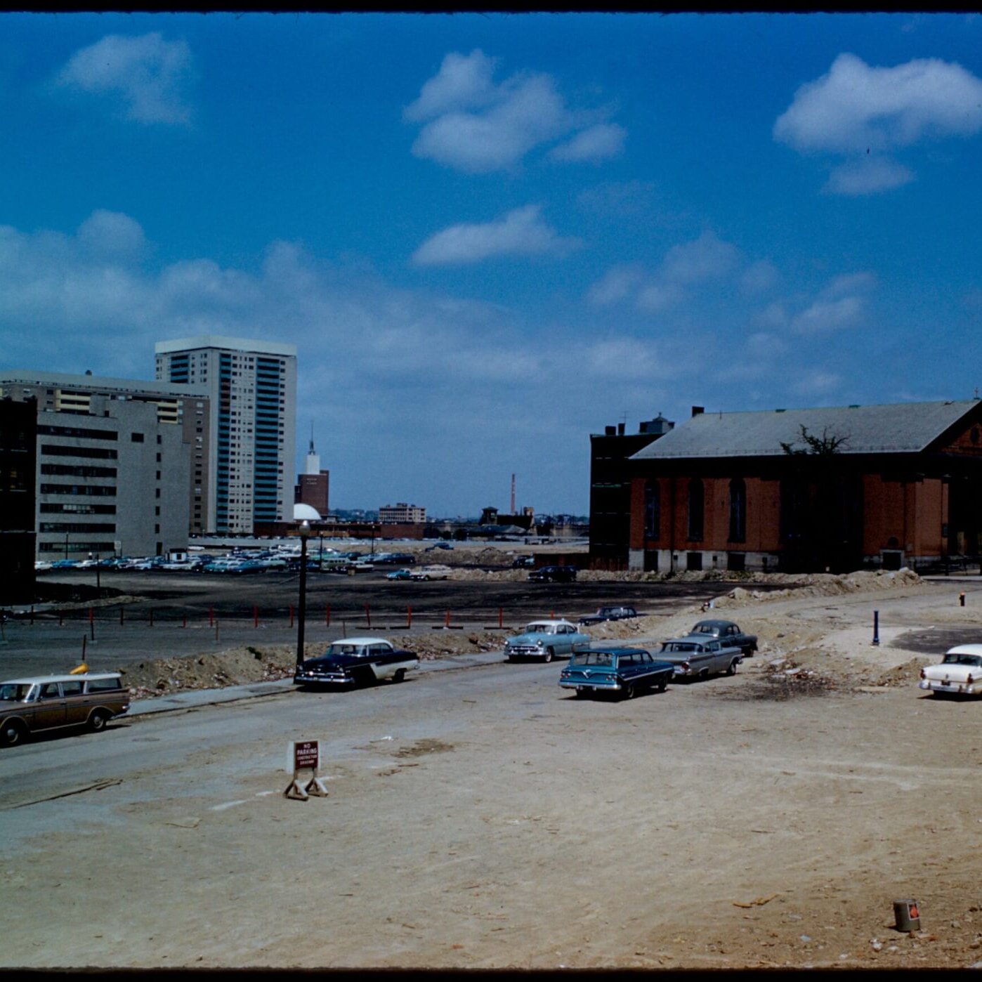 A mostly empty area with many parked cars with one brick church and two high rises.
