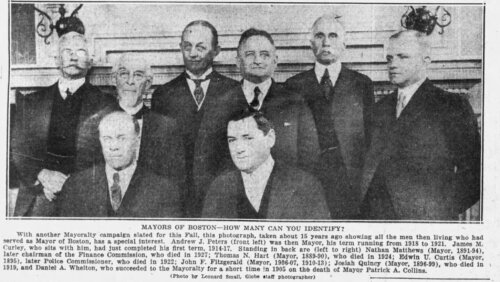 Eight men in suits standing or sitting in front of a marble fireplace.