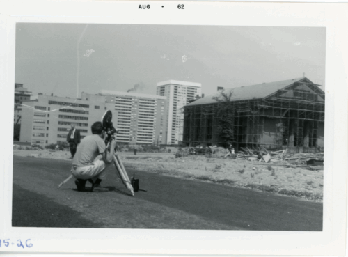 A man crouches behind a TV camera as it faces a church with scaffolding in an empty lot.
