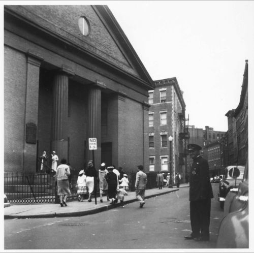 A group of parishioners climbing the steps of a church while a policeman looks on.