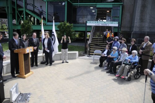A group of people in wheelchairs facing a man and a few others standing behind a podium. They are sitting in front of a transit station.