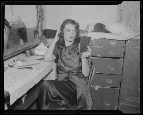 A woman sitting at a dressing room vanity looking at herself in a mirror.