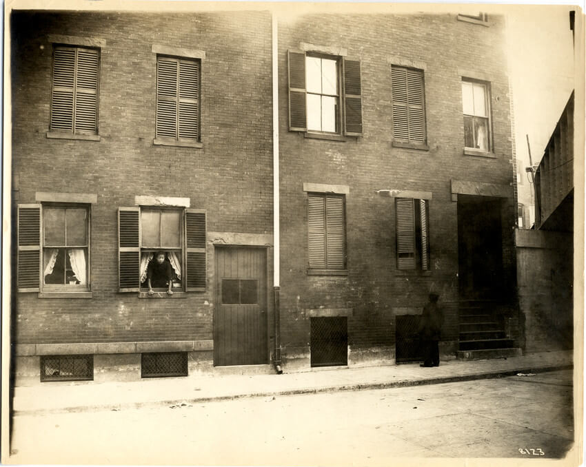 Two tenement buildings with a woman looking out a curtained window.