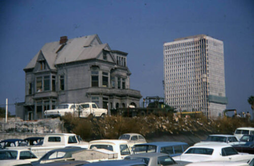 An old Victorian mansion next to a parking lot with a large high rise behind it.
