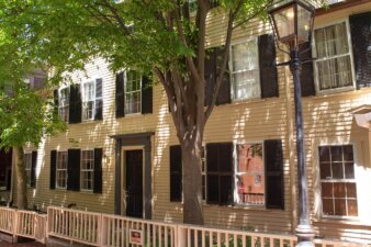 A yellow federalist style house with black shutters and two trees in front.