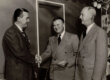 Three men standing with two shaking hands.