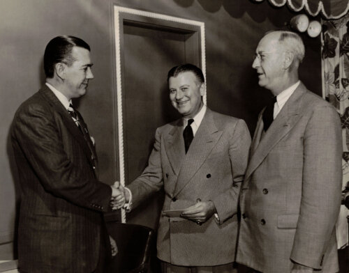 Three men standing with two shaking hands.