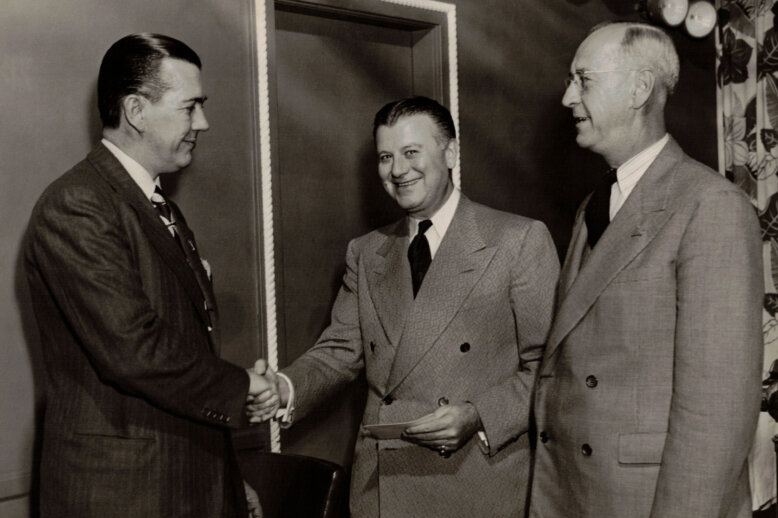Three men standing with two shaking hands.