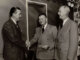 Three men standing with two shaking hands.