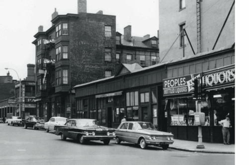 A black and white photo of a street corner with cars and shops.