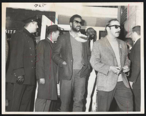 Two men in sunglasses escorted out of a building by several men in suits and a police officer.