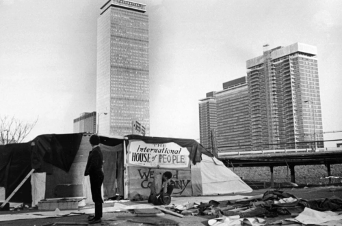 A person stands in front of a tent in a parking lot with sky scrapers in the distance.
