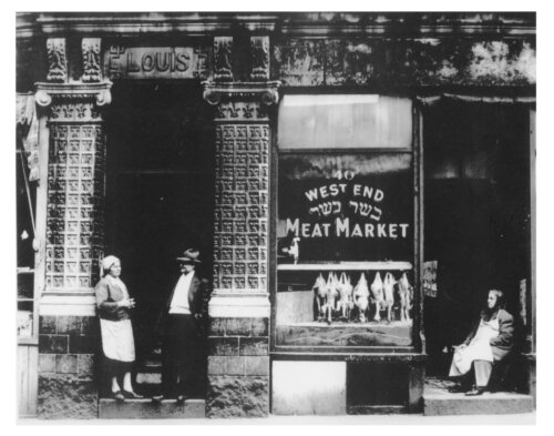 Three people standing in the doorways of a meat market with birds hung in the window.
