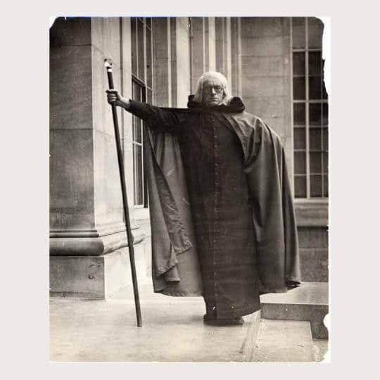 A man with white hair dressed in wizard robes with a staff poses in front of a building.