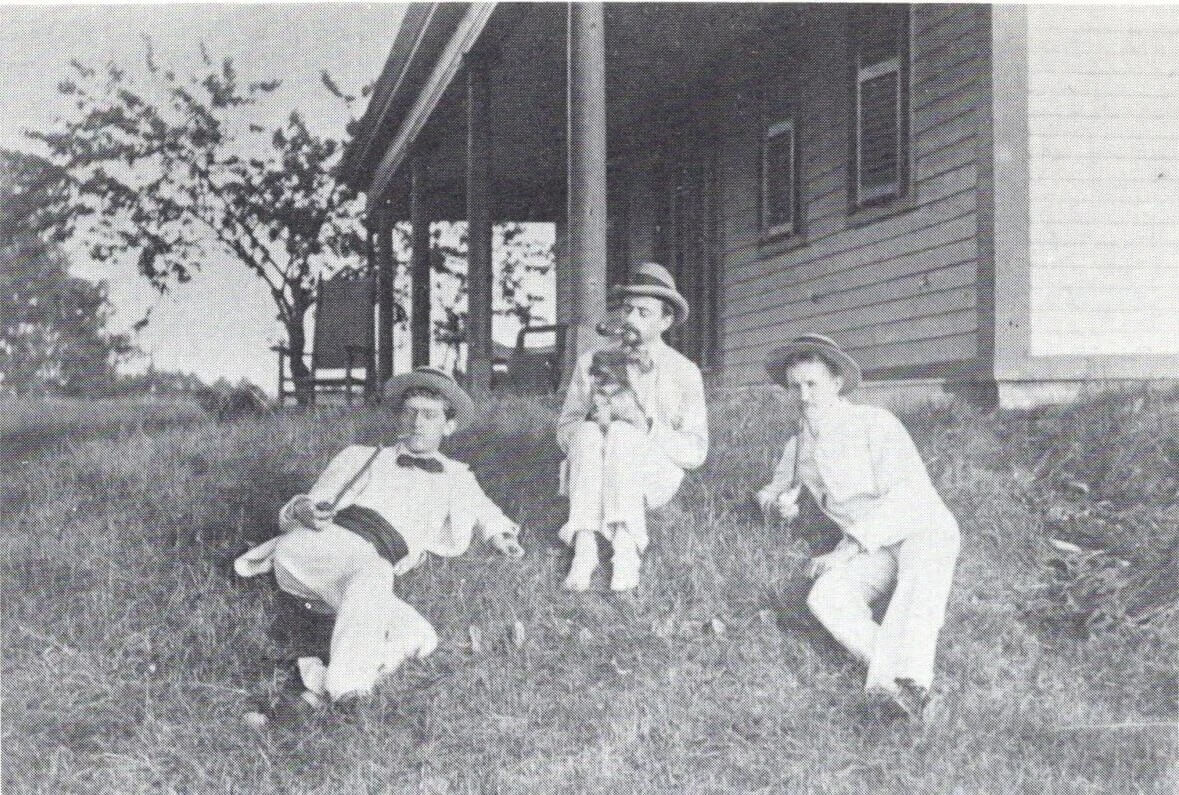 Three men in white suits lie on a grassy hill in front of a low porch.
