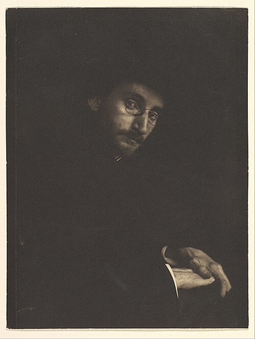 A mostly dark photo of a man in round glasses staring at the camera.