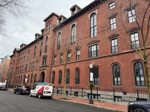 The side of a brick building with many windows and a metal fence around the outside.