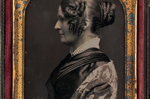 A portrait of a woman facing to the left with a bun and elaborate curls at the side of her face.