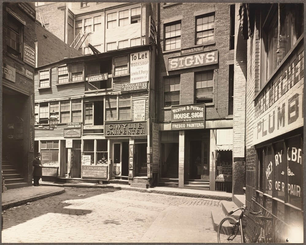 A narrow cobbled street at with many small business signs on the side of the buildings.