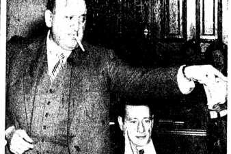 A man in a suit with a cigarette in his mouth pointing at someone off camera. Another man sits next to him.