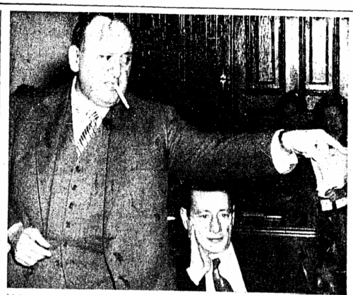 A man in a suit with a cigarette in his mouth pointing at someone off camera. Another man sits next to him.