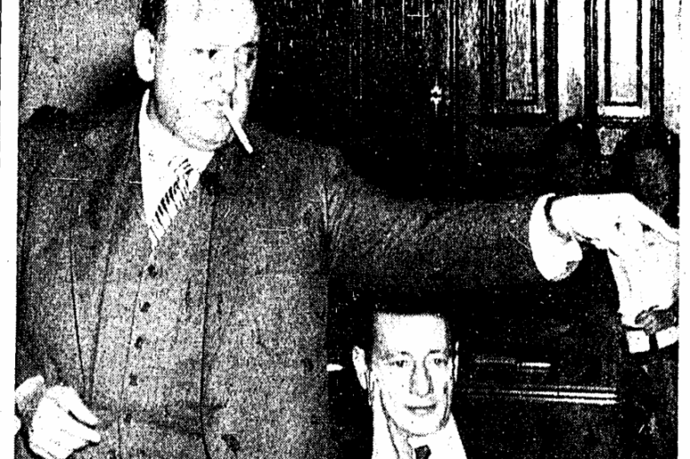 A man in a suit with a cigarette in his mouth pointing at someone off camera. Another man sits next to him.