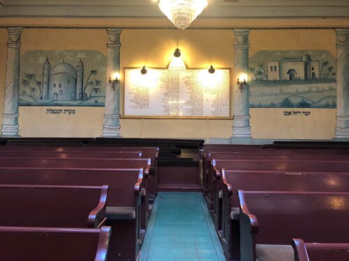 A raked seating section in a sanctuary with pews leading back towards two murals of stone buildings.