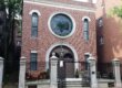 The front facade of a building with a large cast iron gate in front of it. The building is made of brick and has a large stained glass window with a Star of David in the center.