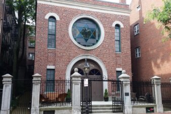 The front facade of a building with a large cast iron gate in front of it. The building is made of brick and has a large stained glass window with a Star of David in the center.