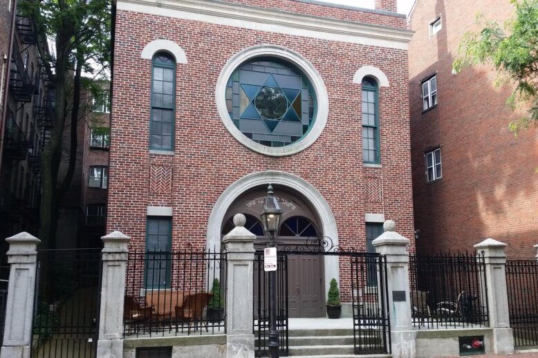 The front facade of a building with a large cast iron gate in front of it. The building is made of brick and has a large stained glass window with a Star of David in the center.