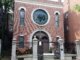 The front facade of a building with a large cast iron gate in front of it. The building is made of brick and has a large stained glass window with a Star of David in the center.