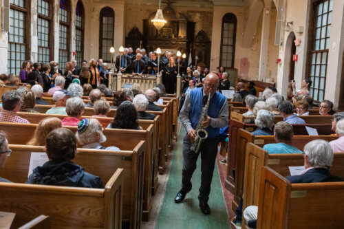 A man playing a saxophone walks down the aisle between two rows of pews.