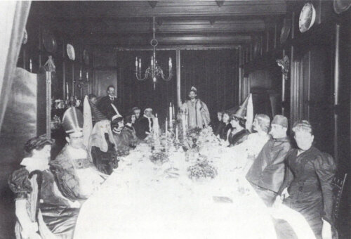 A group of men and women sit in a formal dining room in medieval costumes in front of an elaborate table setting.
