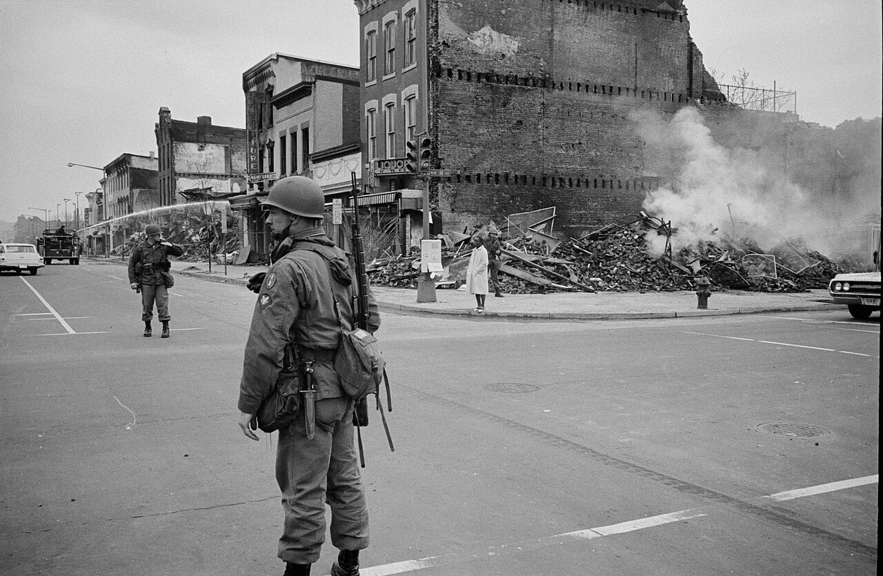 A soldier stands in a street intersection while a building is burning on the corner and a fire truck tries to put it out.